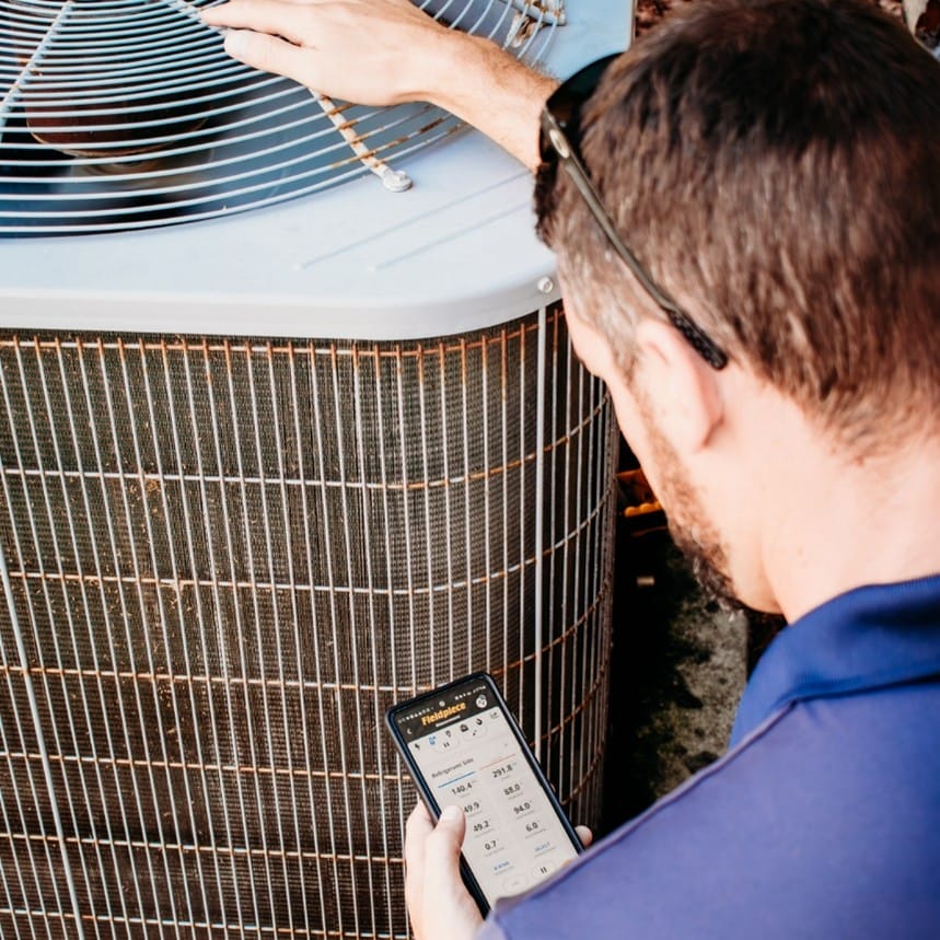 HVAC technician checks an outdoor AC condenser while reading system performance data on a smartphone during a service call.